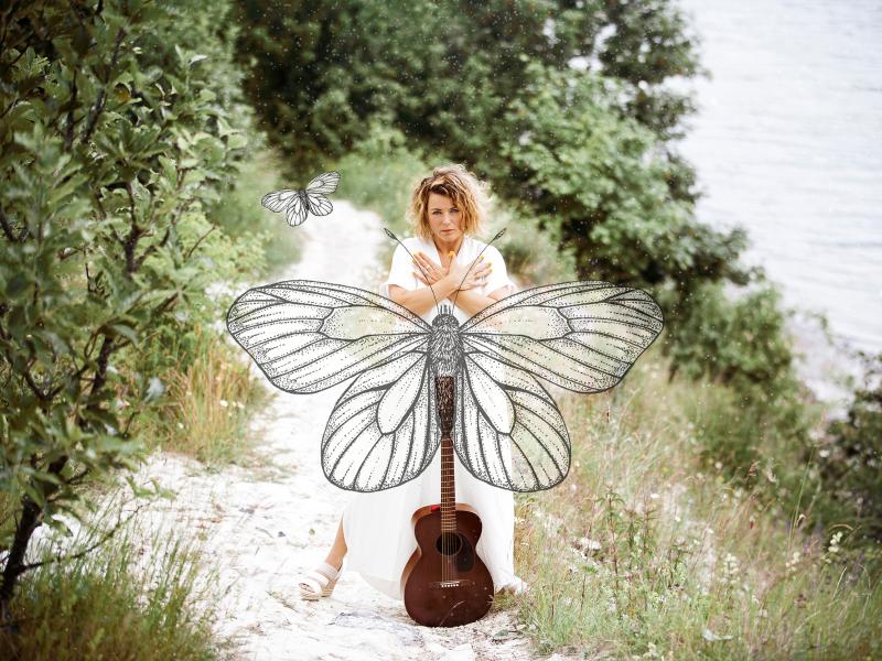 Billede af Stine Michel i naturen med en stor sommerfugl og en guitar. Fotograf: Signe Find Larsen.